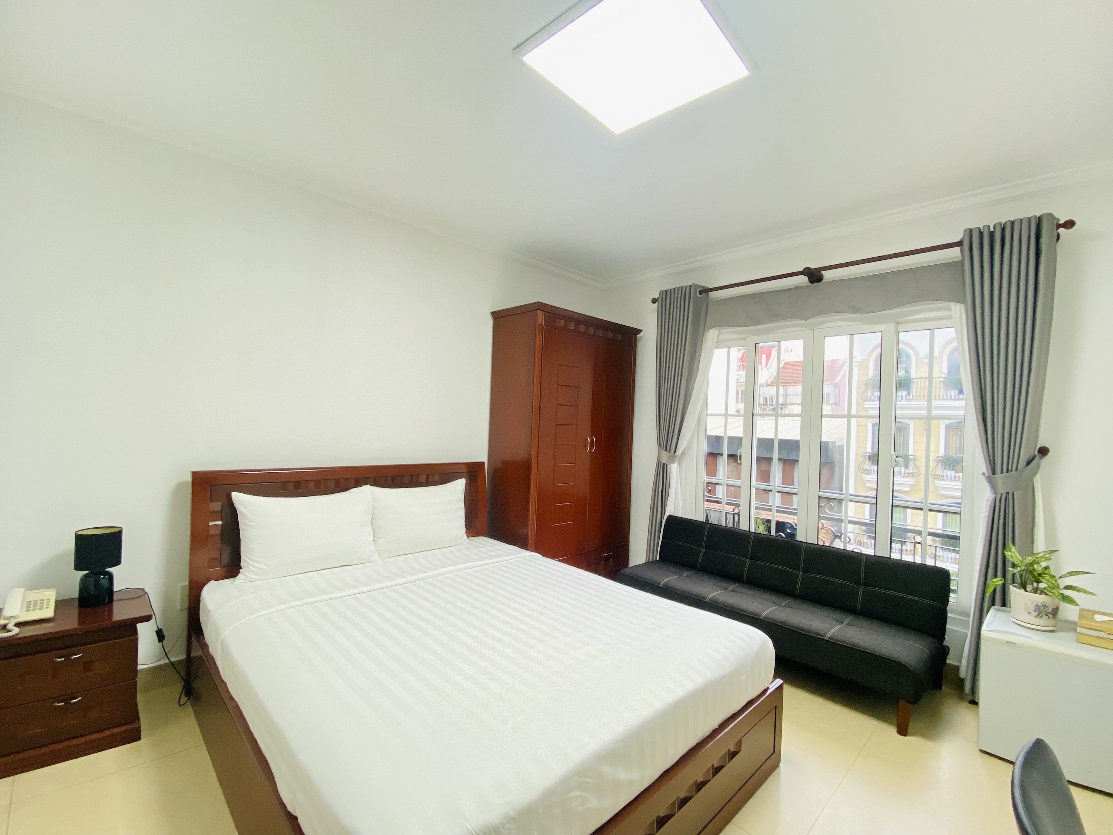 A hotel room with a bed, a wooden wardrobe, a sofa, a desk with a chair, and a window with curtains.
