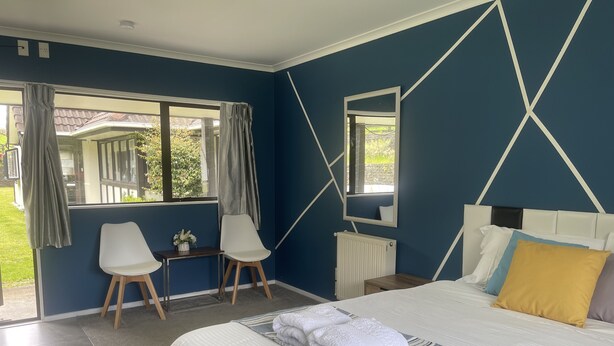 A bedroom with a large bed, a small table with a vase, two chairs, and a window with curtains. - Westminster Mount View Unit 1 (Rotorua)