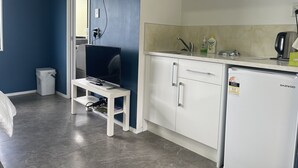 A compact kitchen with a white refrigerator, a sink, and a microwave. There is a small table with a chair and a trash bin. - Westminster Mount View Unit 1 (Rotorua)