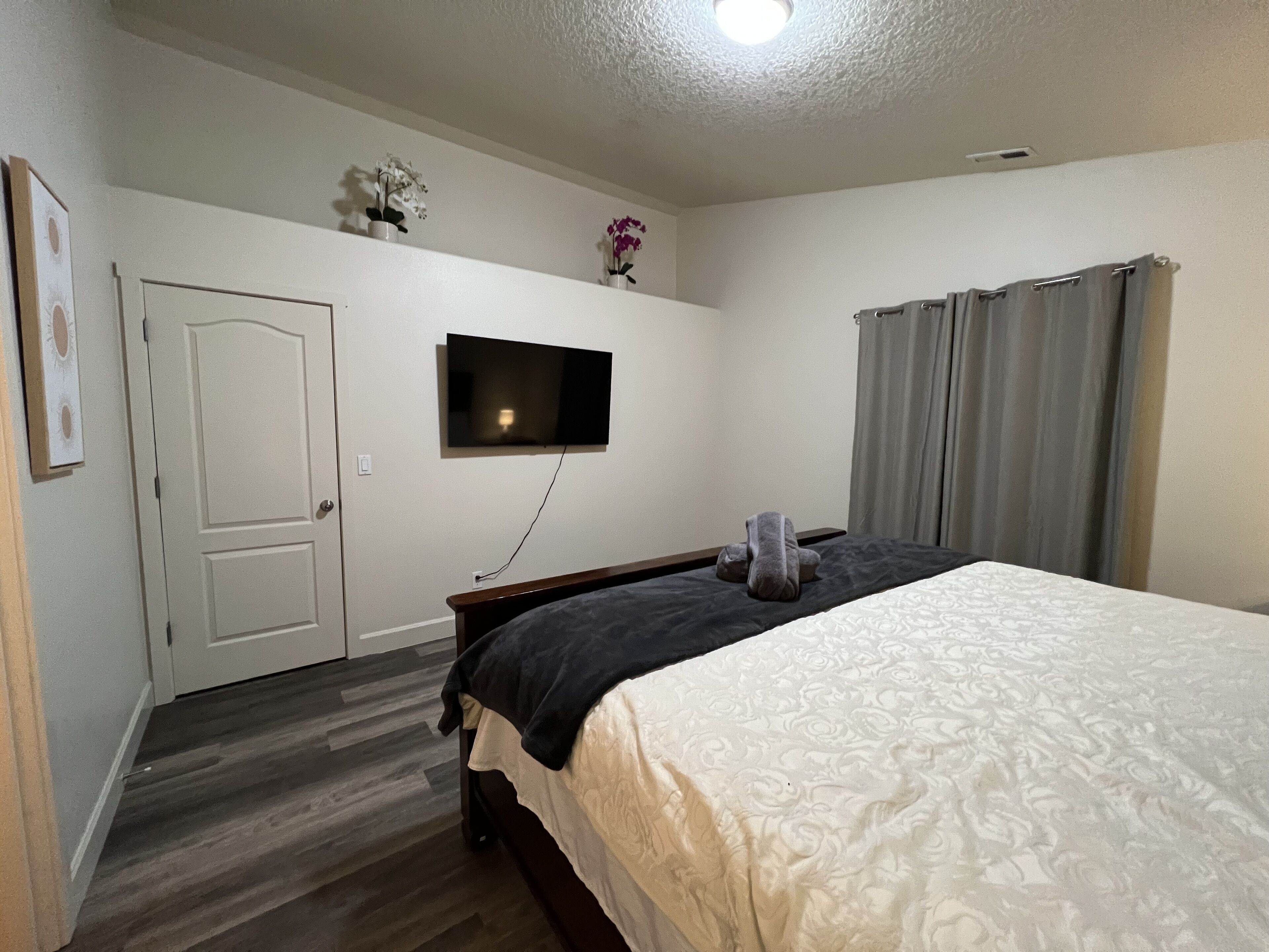 A bedroom with a bed, a flat-screen TV mounted on the wall, a door, and a window with curtains.