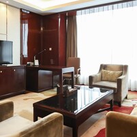 Habitación de hotel con un televisor de pantalla plana grande, un escritorio de madera, un sillón de cuero, un sofá y una mesa de centro de vidrio.
