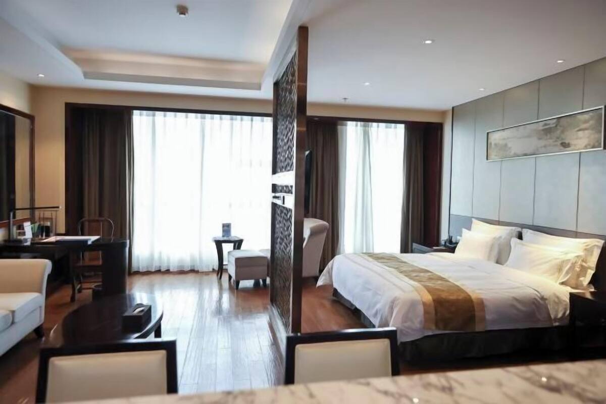 Ein Hotelzimmer mit einem großen Bett, einem Schreibtisch, einem Sessel und Blick ins Freie durch das Fenster.