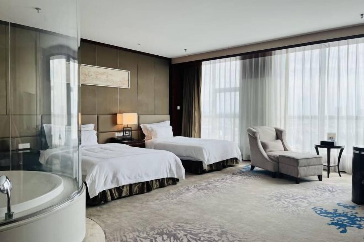 Ein Hotelzimmer mit zwei Betten, einem großen Fenster mit durchsichtigen Vorhängen, einer Sitzecke mit einem Sessel und einer Badewanne.