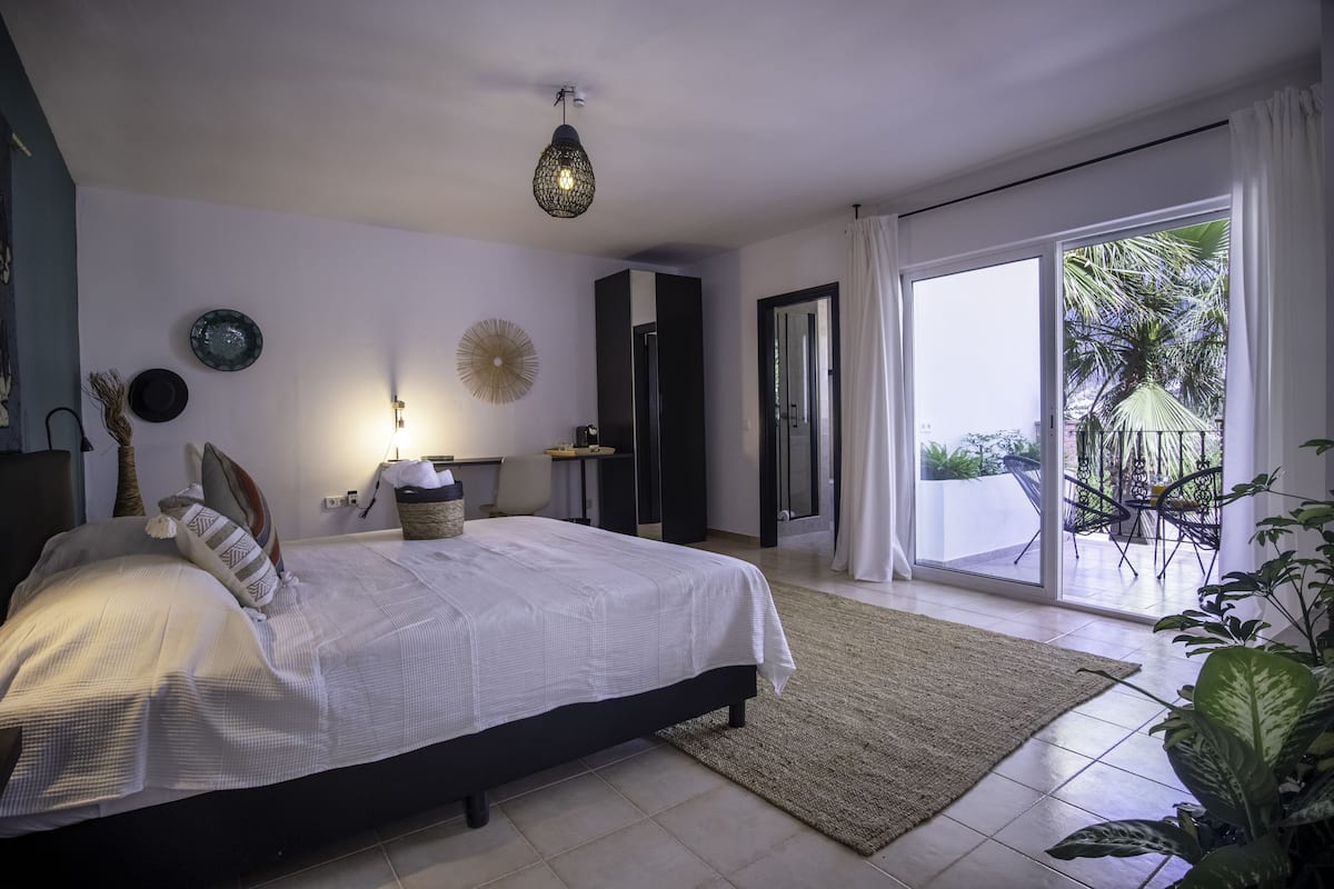 Une chambre à coucher comprenant un lit, une table de chevet, une lampe, un miroir et donnant sur le balcon extérieur.