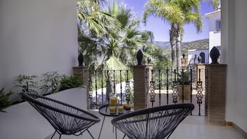 Un balcón con dos sillas de alambre negras, una mesita con bebidas y vistas a palmeras y un edificio al fondo.