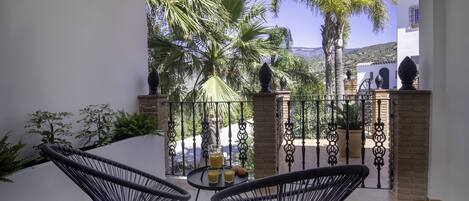 A balcony with two black wire chairs, a small table with drinks, and a view of palm trees and a building in the background.