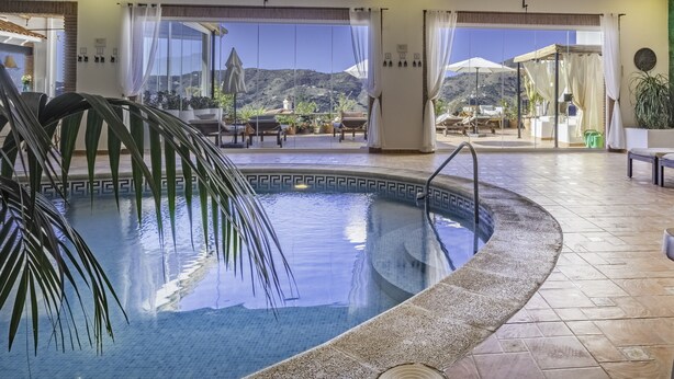 An indoor swimming pool with clear views of the outdoors, including a patio area with lounge chairs and umbrellas. - Malaga Hills, Adults Only boutique, wellness & hiking Eco-Hotel (Còmpeta)
