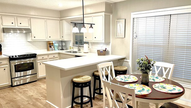 Una cocina moderna con armarios blancos, una isla central, taburetes de bar y una mesa de comedor con un mantel individual colorido.