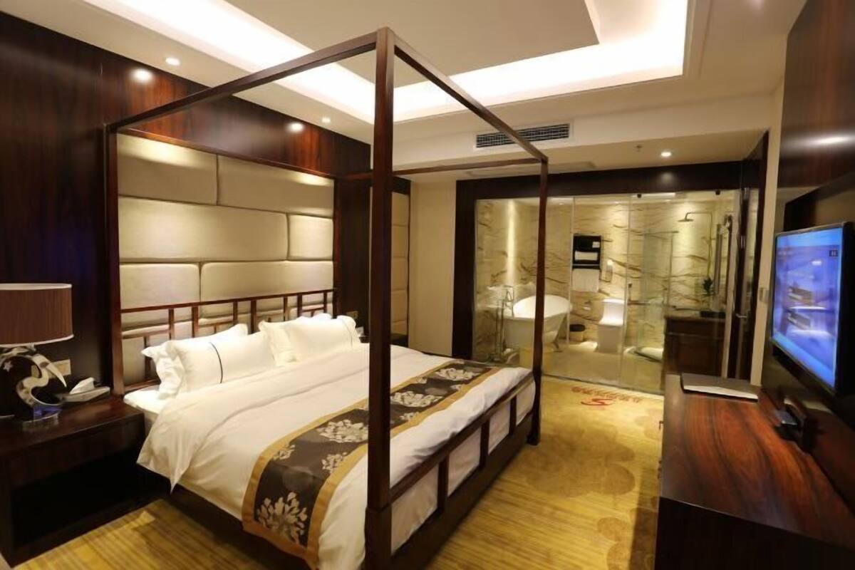 A four-poster bed with a wooden frame and white bedding, a television on the right, and a bathroom visible through a glass door.