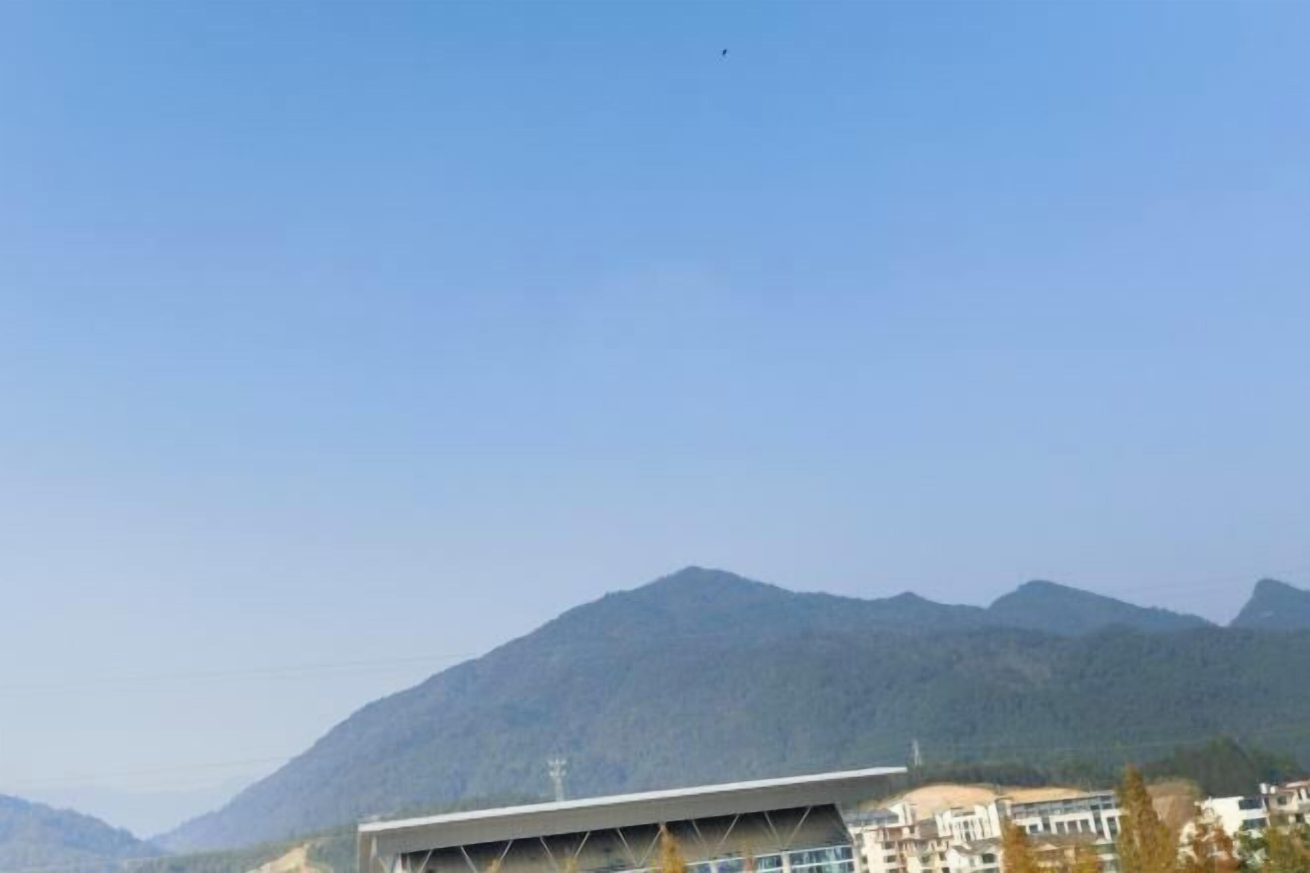 Un edificio moderno con techo plano y amplios ventanales, rodeado de montañas y otros edificios.