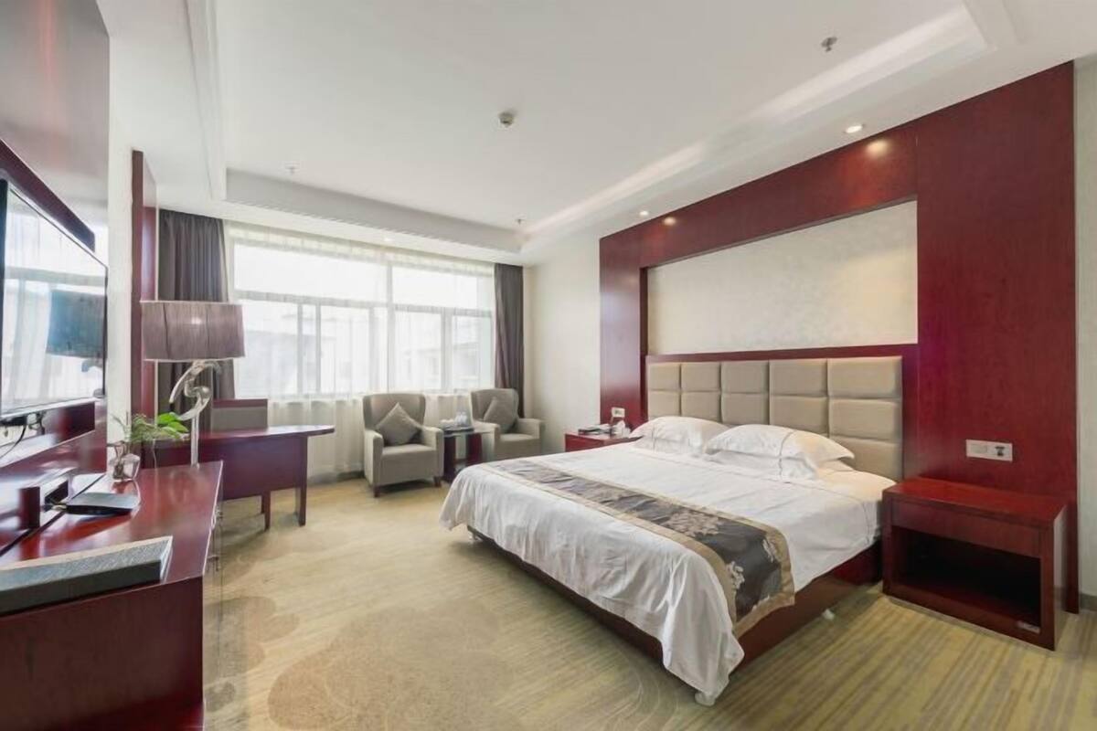 A hotel room with a large bed, a desk with a lamp, two armchairs, a TV, and a large window.