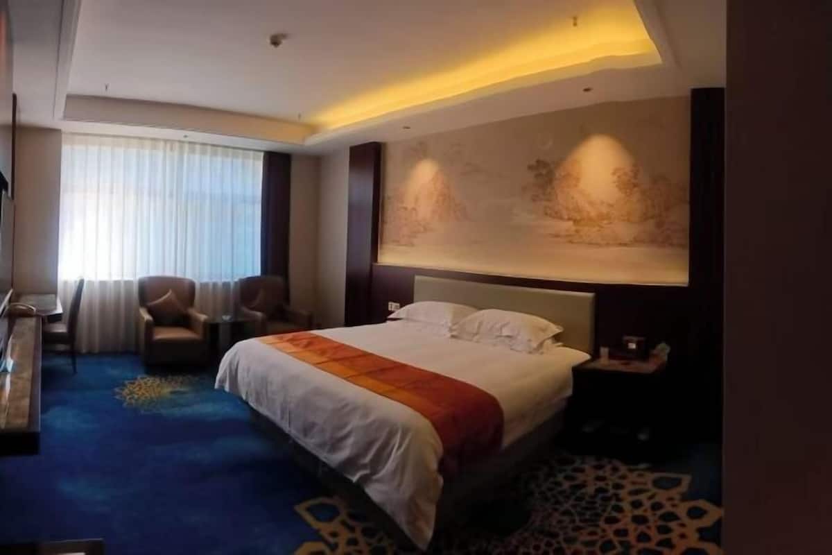 A hotel room with a large bed, a desk with a chair, a small table, and a large window with curtains.