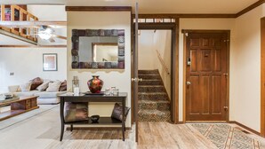 Una sala de estar con sofá, mesa de centro y un espejo decorativo. Una escalera con alfombra estampada lleva a una puerta de madera.