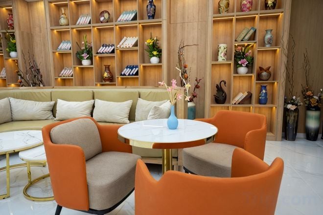 Een moderne woonkamer met een bank, oranje fauteuils, een ronde tafel en een boekenplank vol decoratieve objecten.