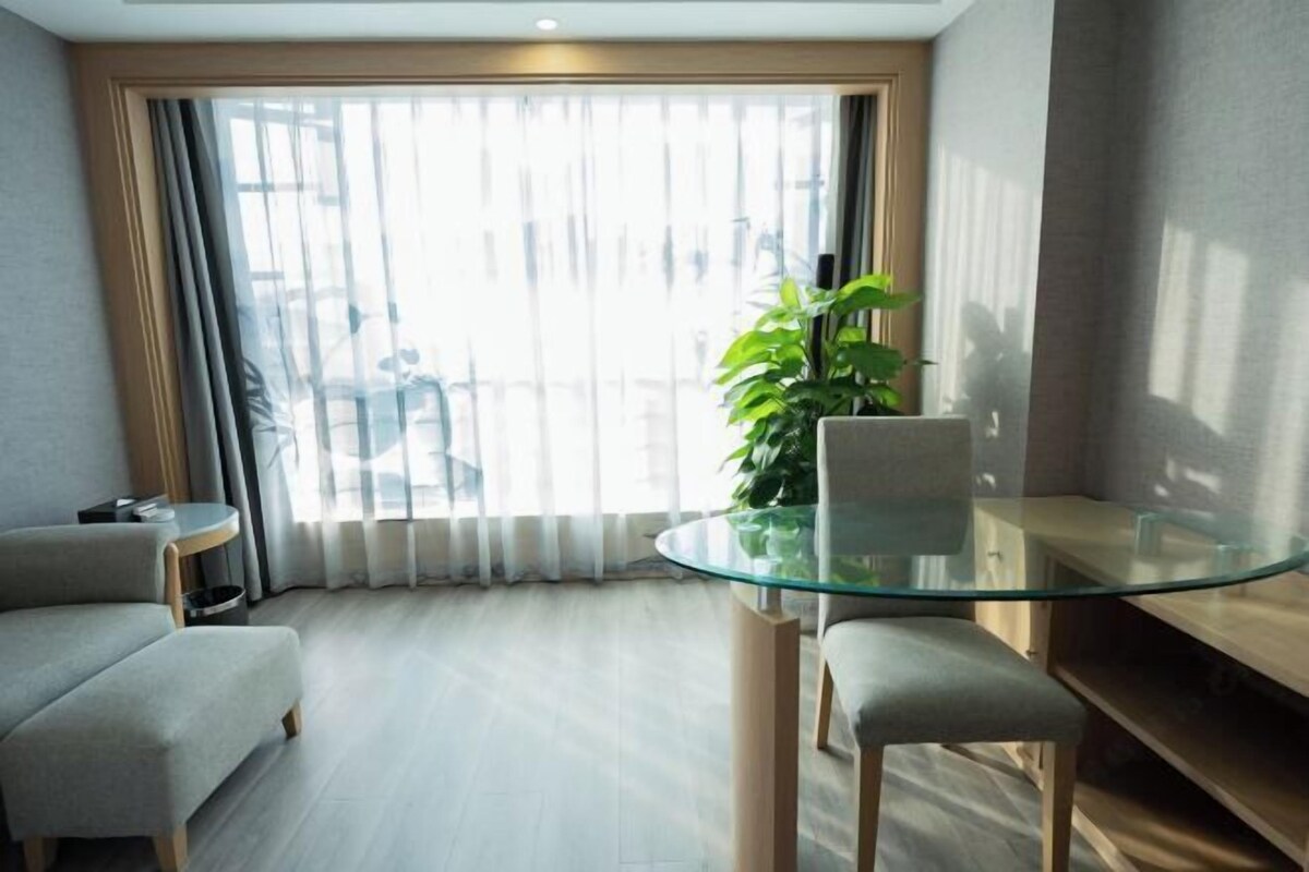 A modern living room with a glass table, a grey sofa, and a large window with sheer curtains.