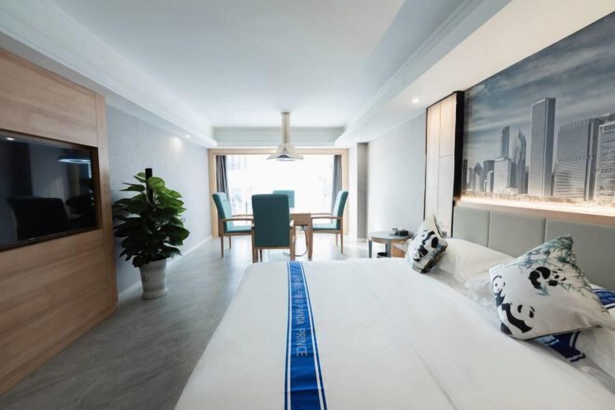 A modern hotel room with a bed, a desk, a chair, a potted plant, and a cityscape painting on the wall.