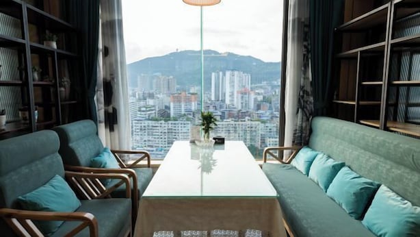 Een moderne woonkamer met een glazen salontafel, twee groene sofa's en houten stoelen. Een groot raam biedt uitzicht op de stad, met gebouwen en bergen op de achtergrond.