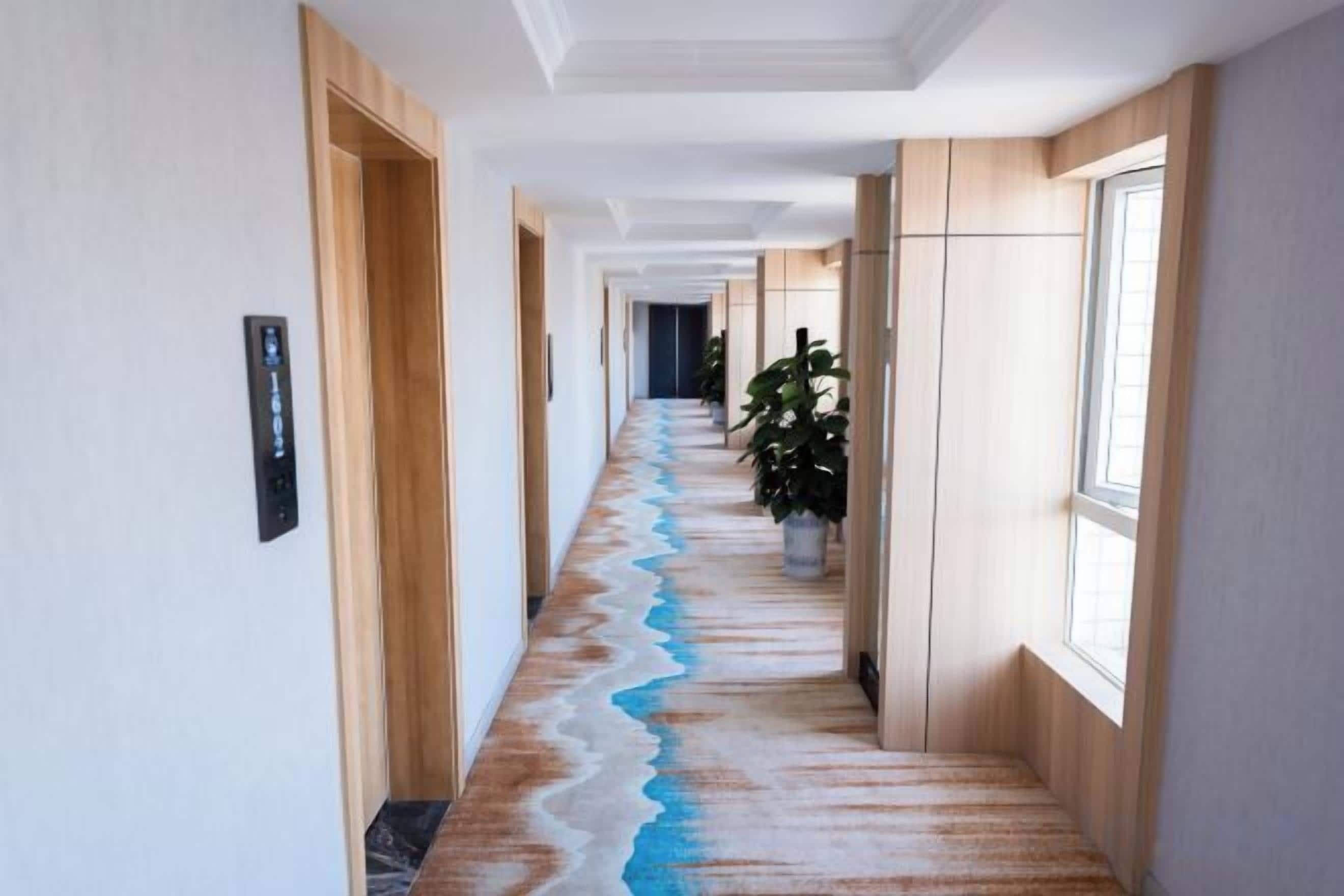 A hotel hallway with a long corridor, wooden doors, a potted plant, and a window on the right.