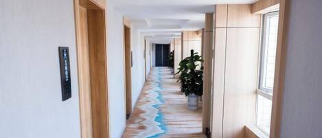 A hotel hallway with a long corridor, wooden doors, a potted plant, and a window on the right.