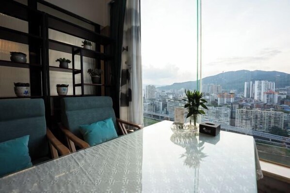 A modern living room with a glass table, teal chairs, a bookshelf with potted plants, and a city view through large windows. - Panda Prince Hotel (Dazhou)