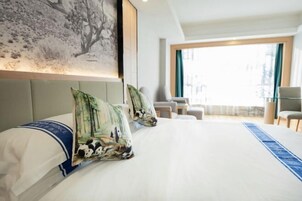 Habitación de hotel con una cama grande, vistas de la ciudad a través de un ventanal amplio y un balcón con mobiliario.