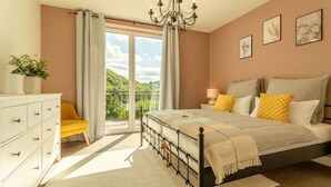 A bedroom with a bed, a dresser, a chair, a balcony with a view of greenery, and a chandelier.