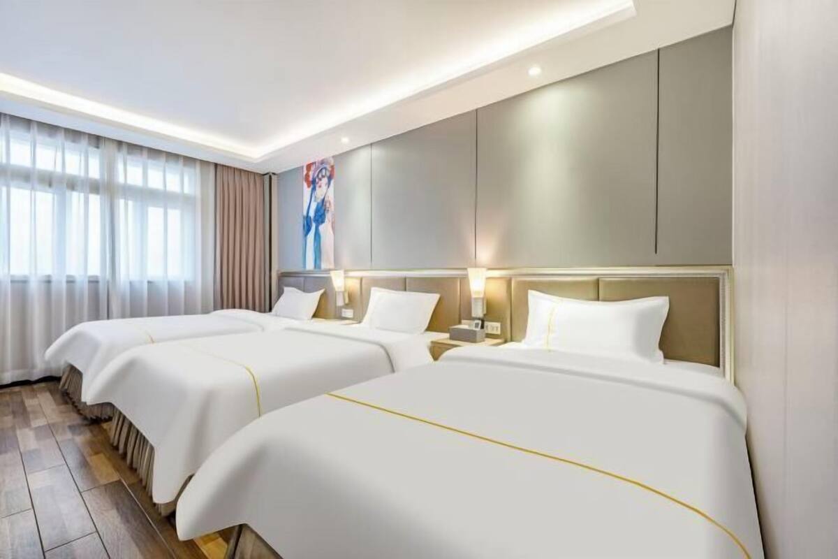 A hotel room with three beds, a large window with curtains, a painting on the wall, and a modern design.