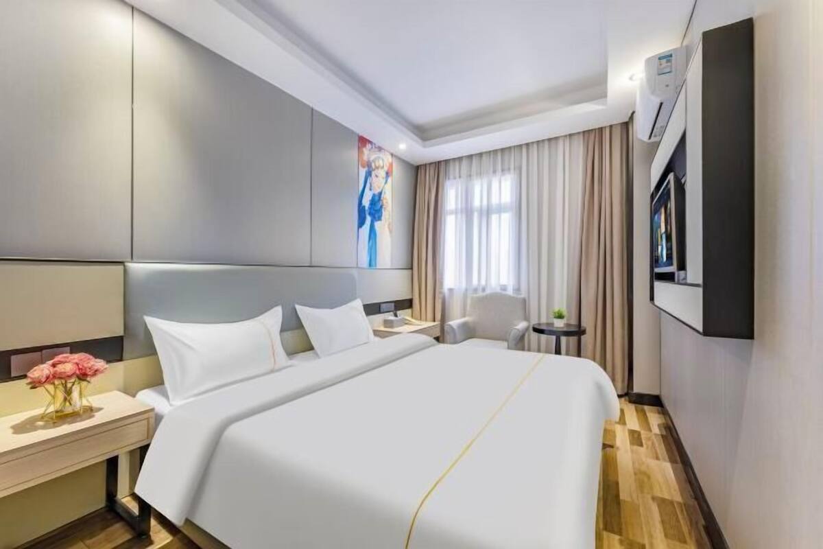 A hotel room with a large bed, a bedside table with a vase of flowers, a small round table with a chair, a TV mounted on the wall, and a window with curtains.