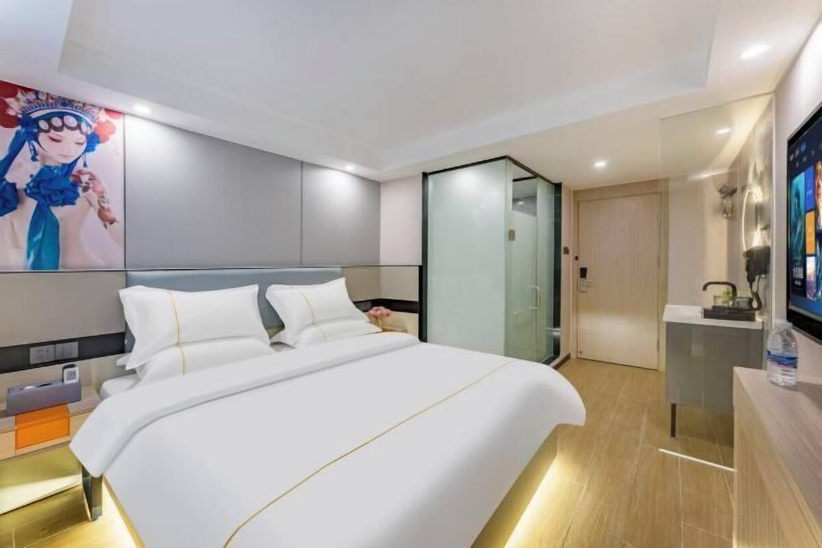 A modern hotel room with a large bed, a glass partition wall, and a flat-screen TV on a wooden console.
