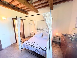 Una cama con dosel y mosquitera, una mesita de noche con lámpara y un aparador con un florero.