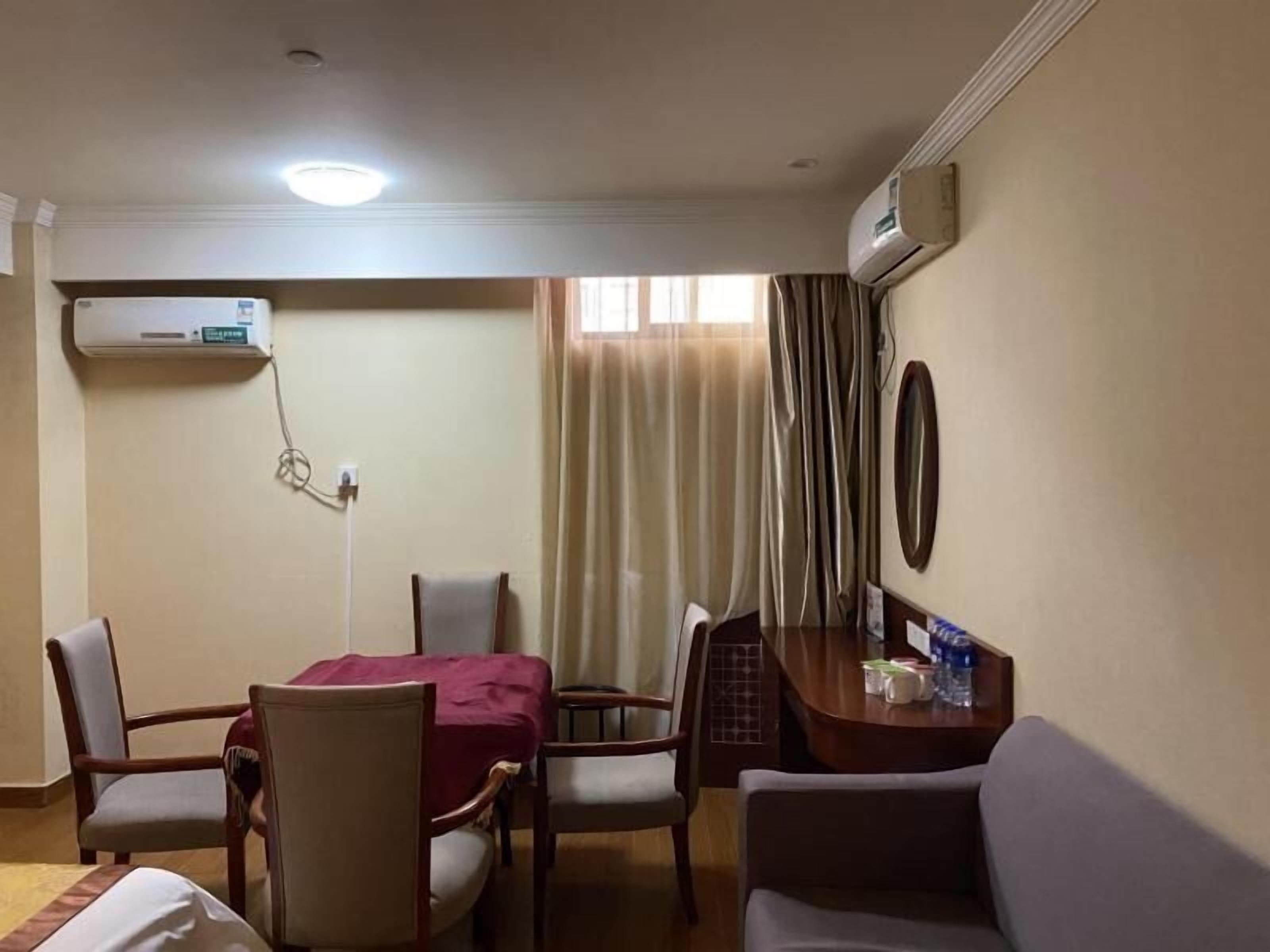 A hotel room with a dining table, two chairs, a sofa, an air conditioning unit, and a window with curtains.