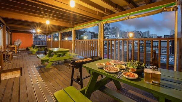An outdoor dining area with wooden tables and benches, a grill, and a view of the city skyline. - Sokcho World Pet-friendly Pension (Sokcho)