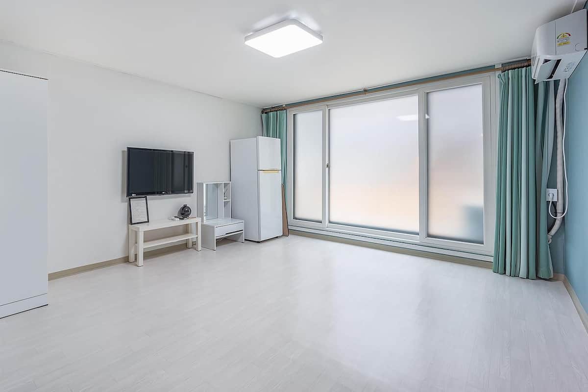 A spacious room with a large sliding glass door, a flat-screen TV, a white wardrobe, and a small desk with a chair.