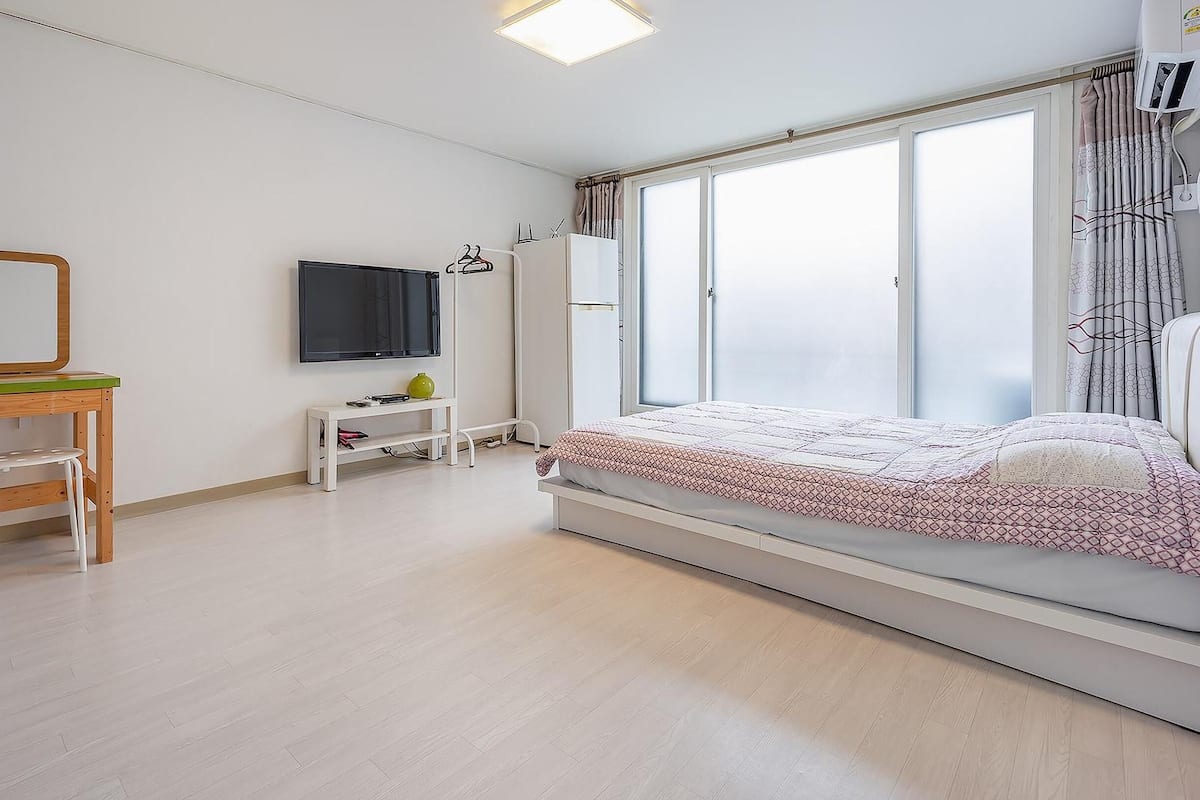 A bedroom with a bed, a desk with a chair, a TV, and a window with curtains.