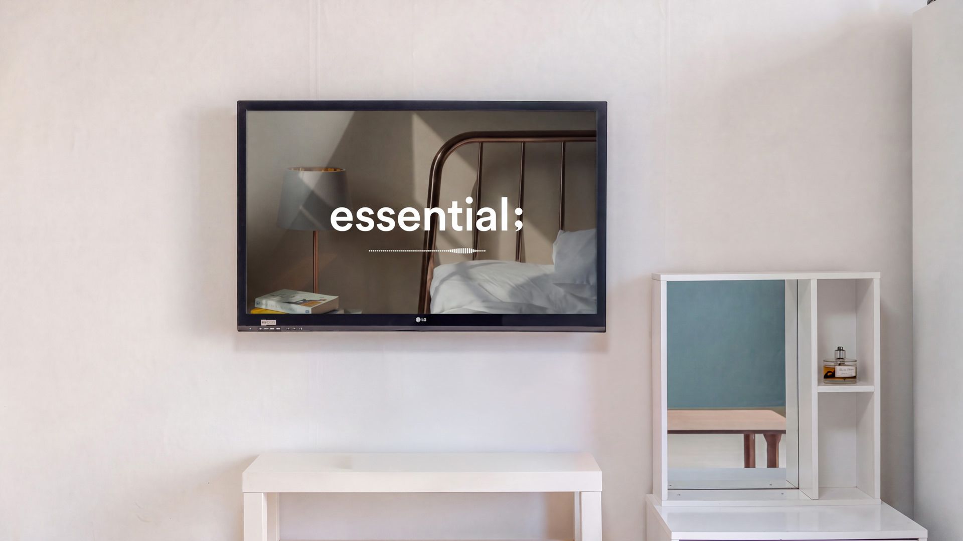 A hotel room with a flat-screen TV mounted on the wall, a bedside table, and a small shelf with a bottle.