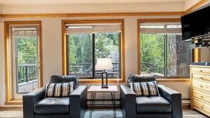 A modern living room with two black leather sofas, a small table, and a lamp. Large windows offer views of a deck and trees. - Slopeside #2775 by Summit County Mountain Retreats (Keystone)