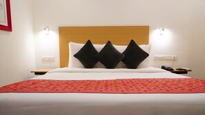 Un letto rifatto con cura, coperto da una trapunta rossa a motivi decorativi, con una testiera in legno e tre cuscini neri. Sono presenti due comodini, uno dei quali con un telefono e un telecomando.
