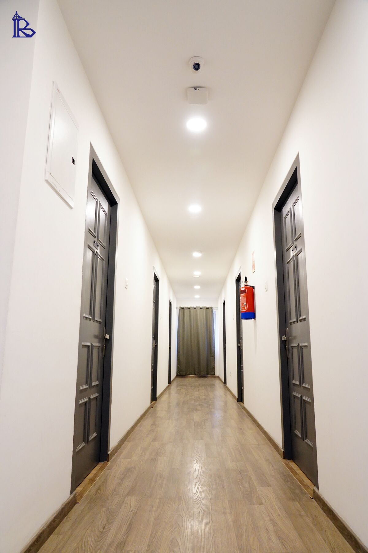Un pasillo largo y estrecho con suelo de madera y paredes blancas. Hay puertas a ambos lados, y un extintor está fijado a la pared.