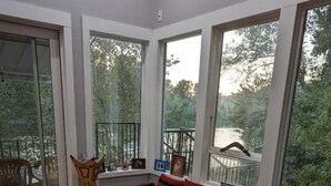 A room with a wooden floor, a red patterned sofa, and large windows offering a view of a forest and a river. - Exotic Waterfront Getaway (Salem)