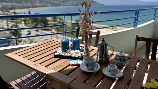 Una mesa de madera con una bandeja de piedras blancas, un jarrón con plumas de plumas, dos velas azules, una tetera y dos tazas en un balcón con vistas al mar.