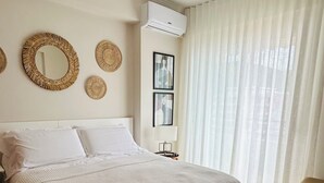Un dormitorio con una cama grande, un ventilador de techo, aire acondicionado instalado en la pared y una ventana con cortinas traslúcidas.
