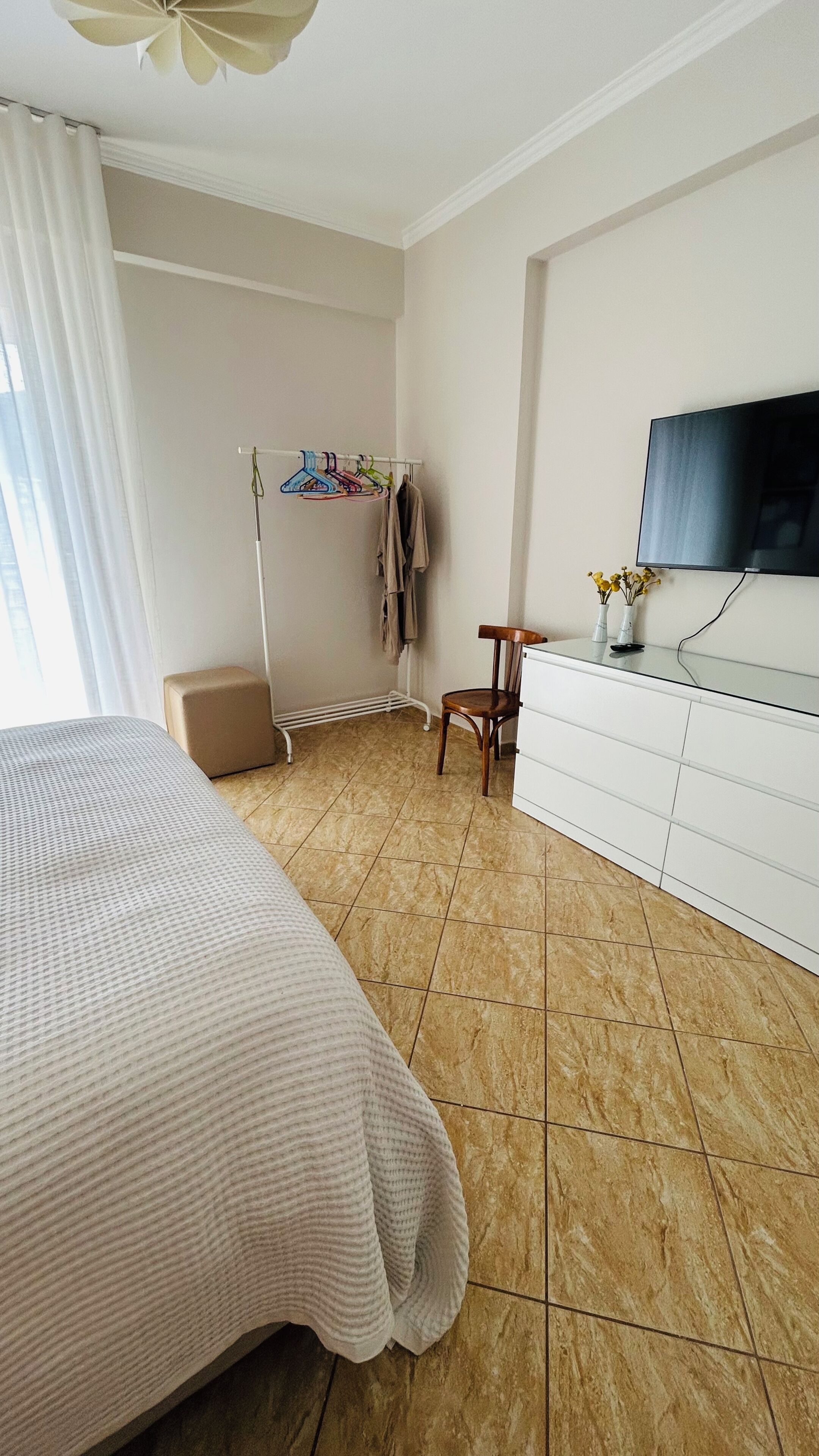 A bedroom with a bed, a chair, a TV, a dresser, and a coat rack with clothes.
