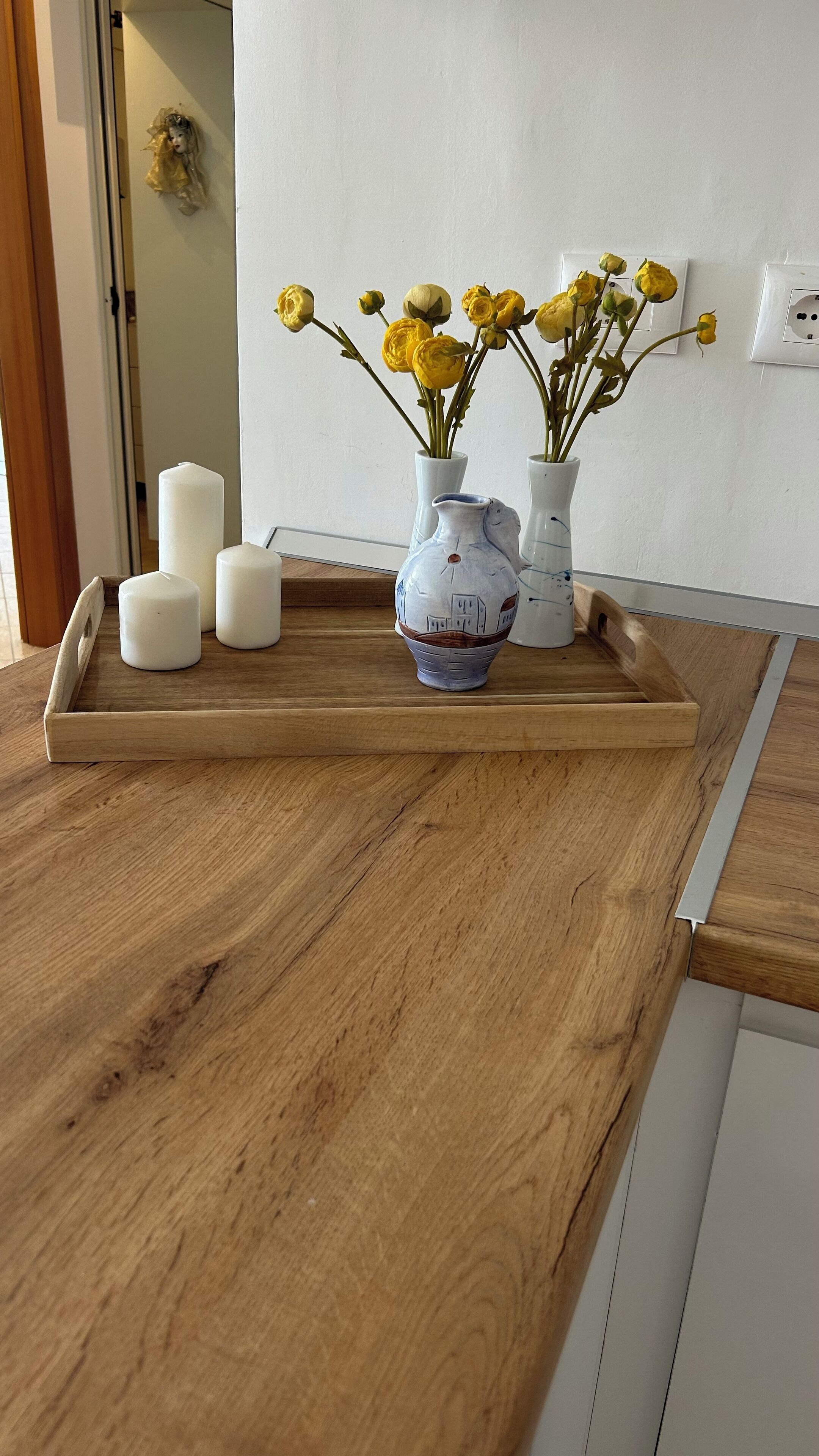 Eine hölzerne Küchenarbeitsplatte mit einer Ablage, auf der drei weiße Kerzen und zwei Keramikvasen mit gelben Blumen stehen.