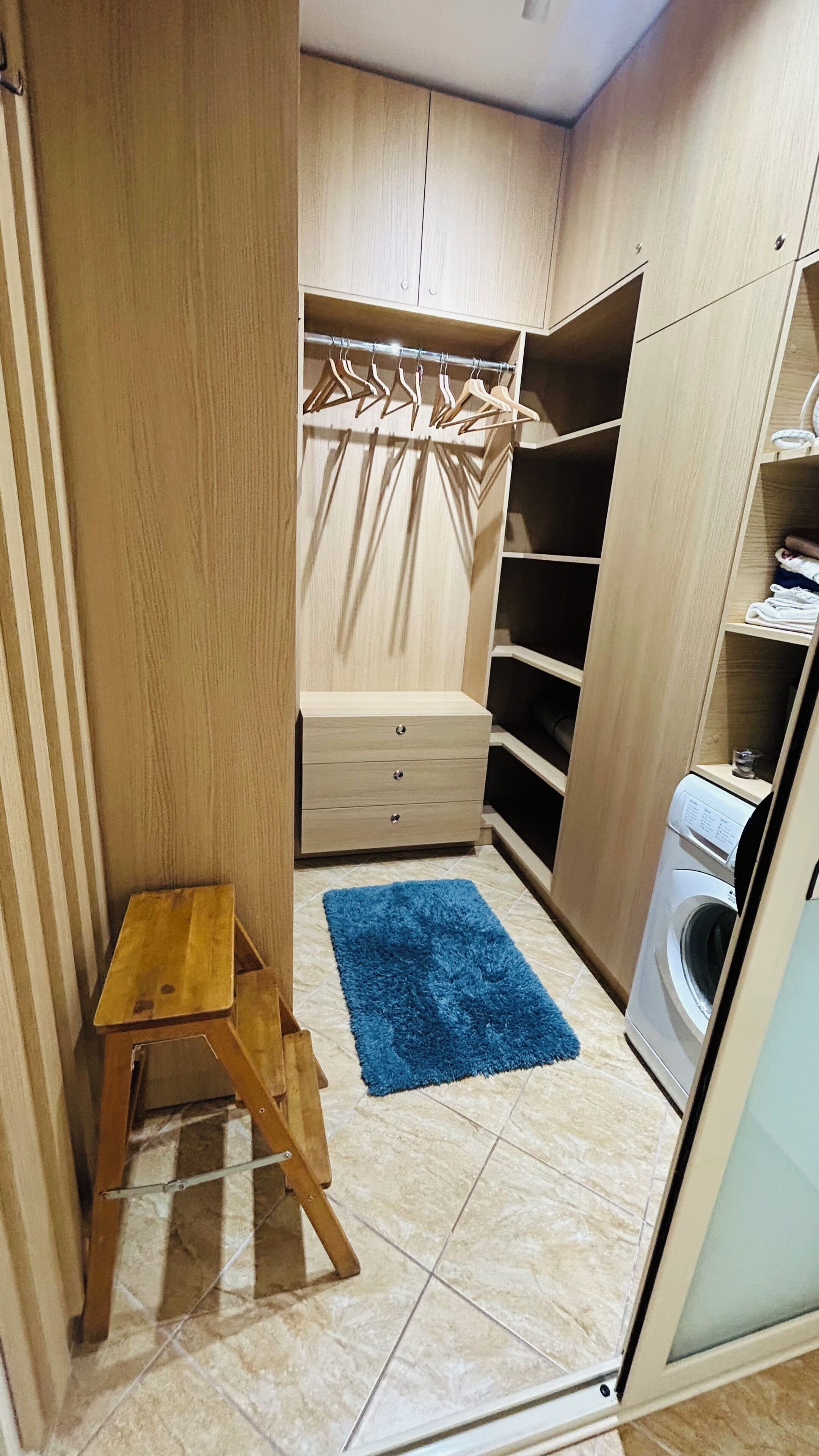 A compact laundry room with a washing machine, a blue rug, a wooden stool, and a clothing rack.