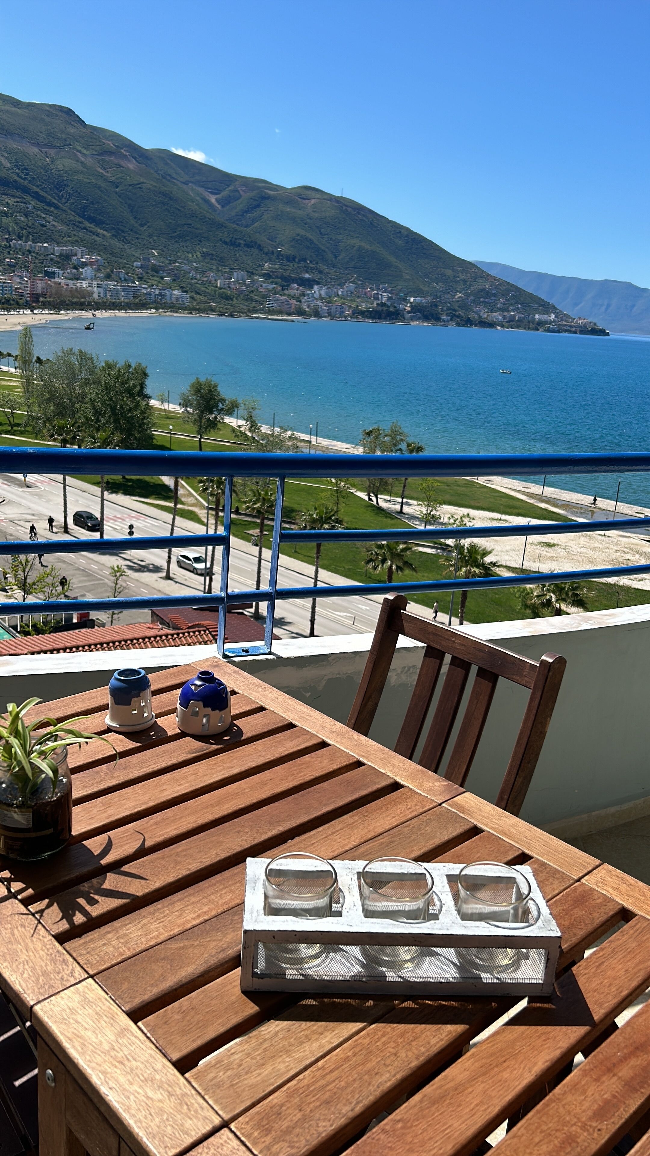 A wooden table with a tray of glasses and a potted plant on a balcony overlooking a coastal view with mountains and a beach.