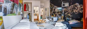 Habitación de hotel con cama, escritorio y zona de estar. Hay un mural en la pared y una ventana con vista a un paisaje urbano.