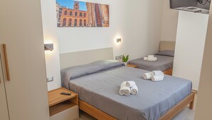 Habitación de hotel con dos camas, cada una con cubrecama gris y toallas blancas. Hay un televisor de pared y un cuadro de un edificio.