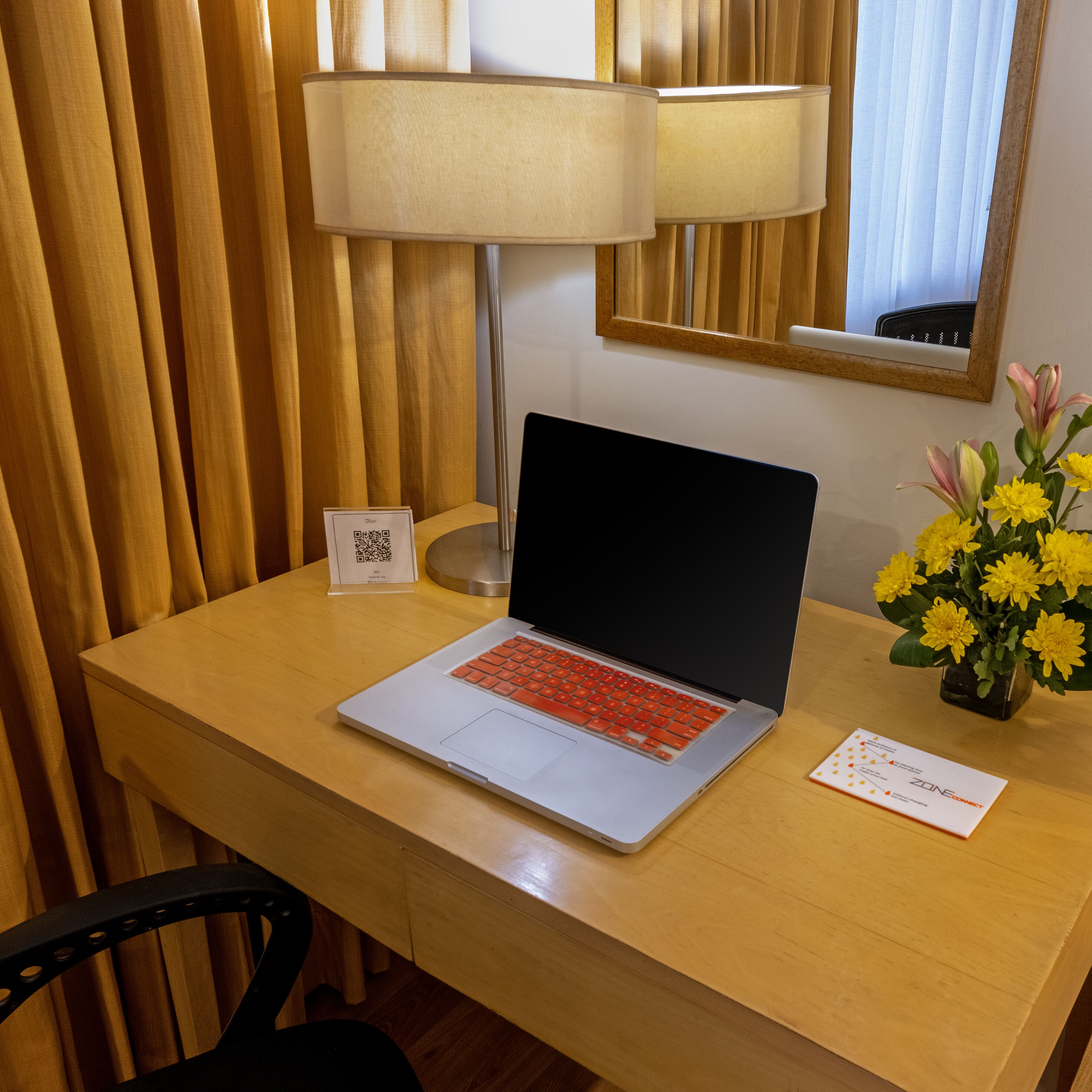 A hotel room desk with a laptop, a lamp, a vase with flowers, and a mirror on the wall.