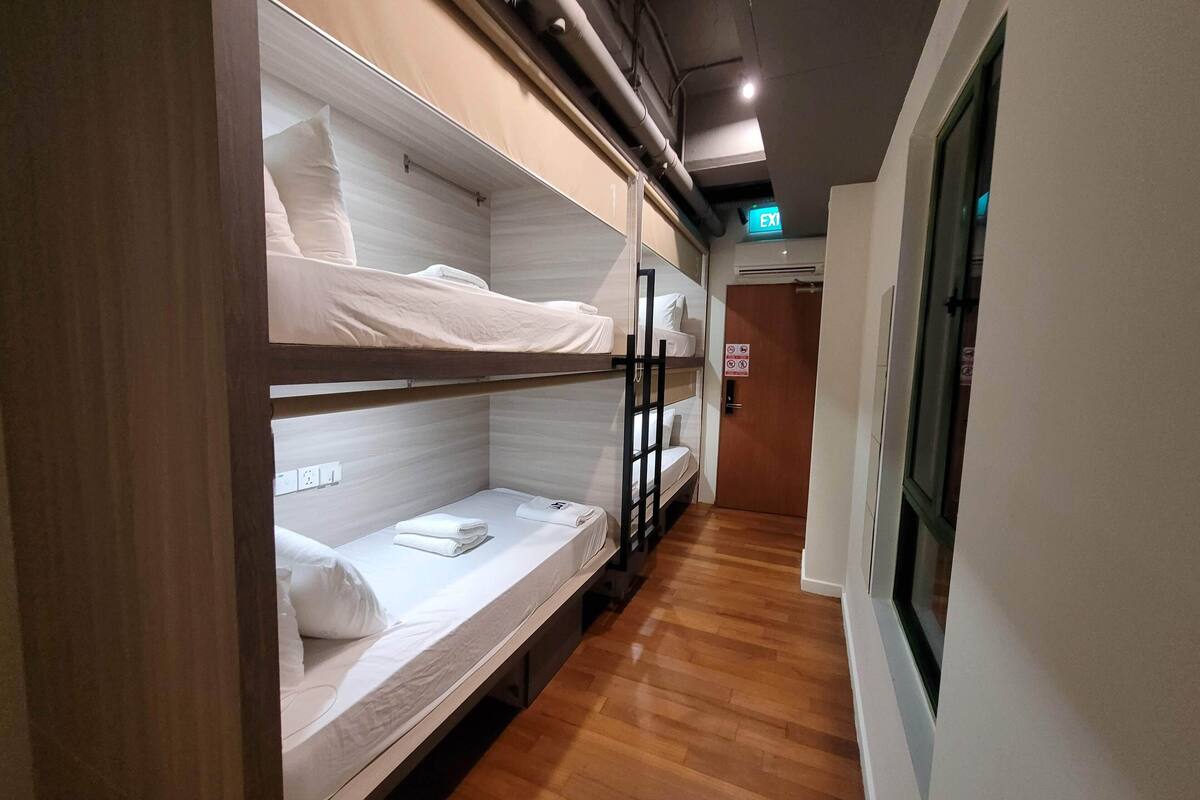 Um corredor com beliches, piso de madeira e uma porta com sinalização de saída.
