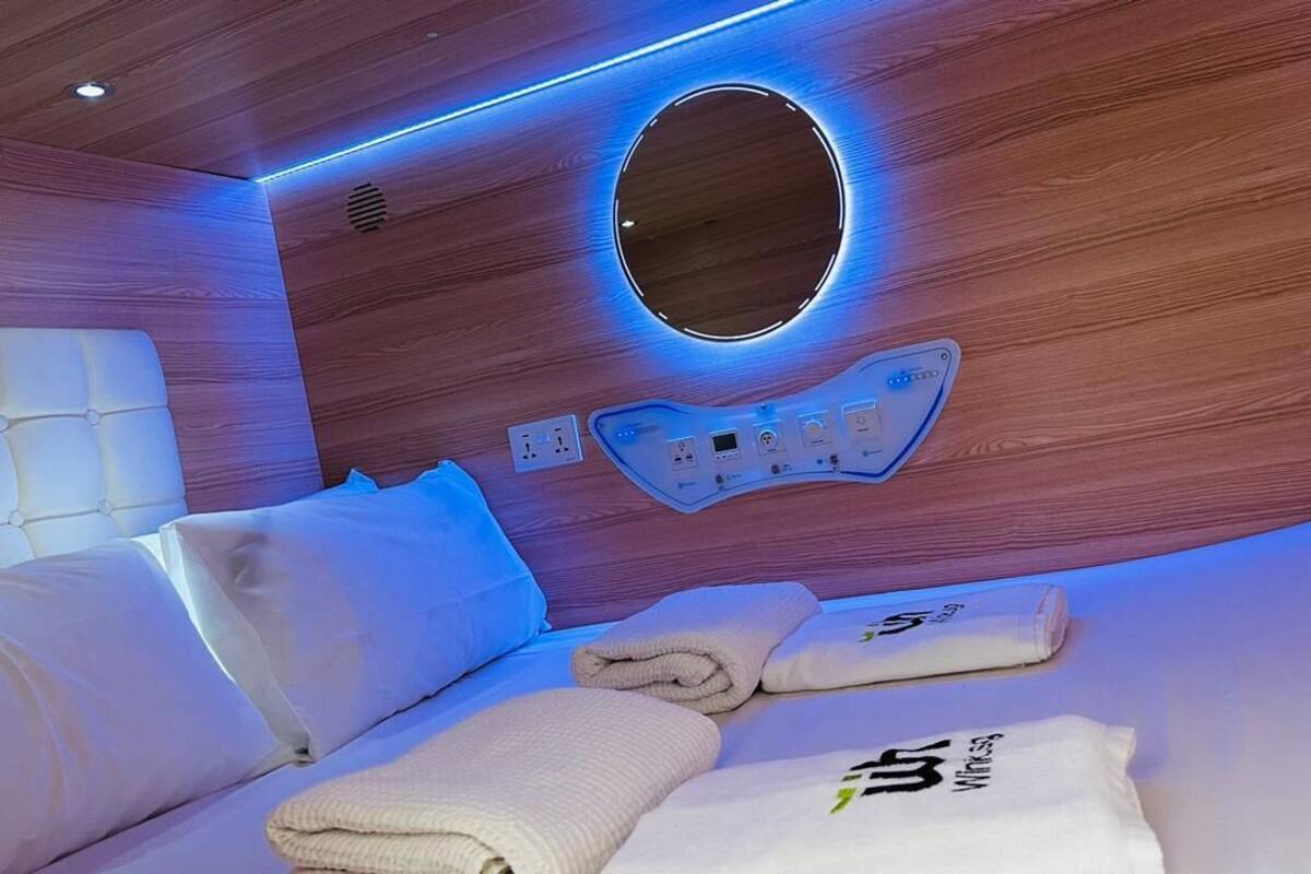 Quarto de hotel moderno com parede de madeira, luminária circular, painel de controle e camas bem arrumadas com toalhas.